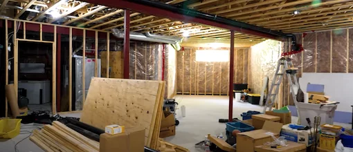 An unfinished basement with exposed wooden framing, insulation, and scattered construction materials, indicating an ongoing renovation project.