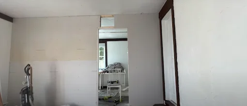 Partially finished drywall installation showing new drywall sheets and wooden framing around a doorway, with a vacuum cleaner nearby.