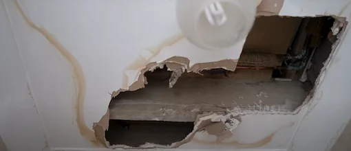 A large hole in drywall with visible water stains surrounding the damage, revealing the structural space behind the wall.